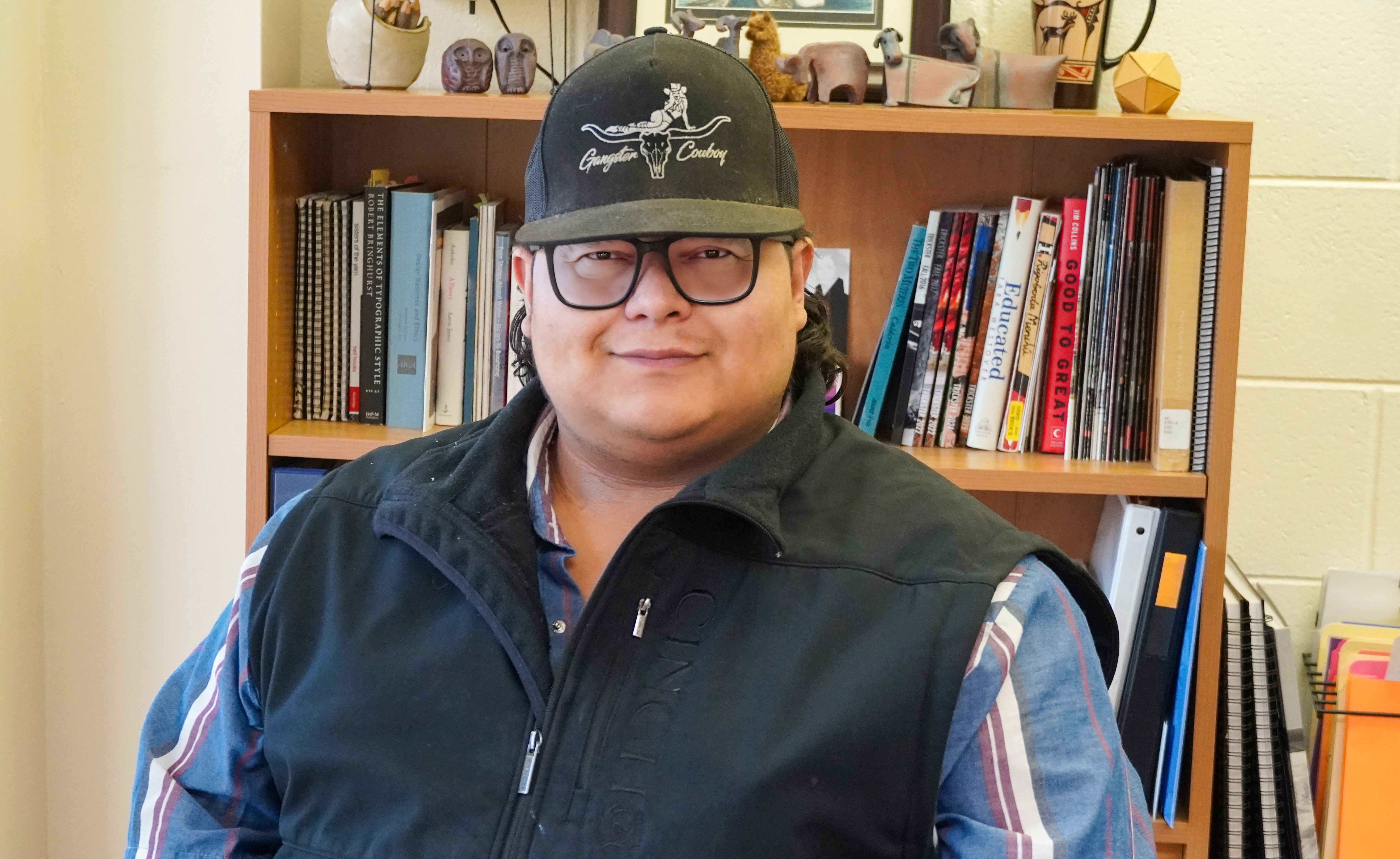 Matt Baldonado wearing dark-rimmed glasses, a baseball cap reading "Gangster Cowboys," a grey vest and a blue shirt with red and white striped accents.